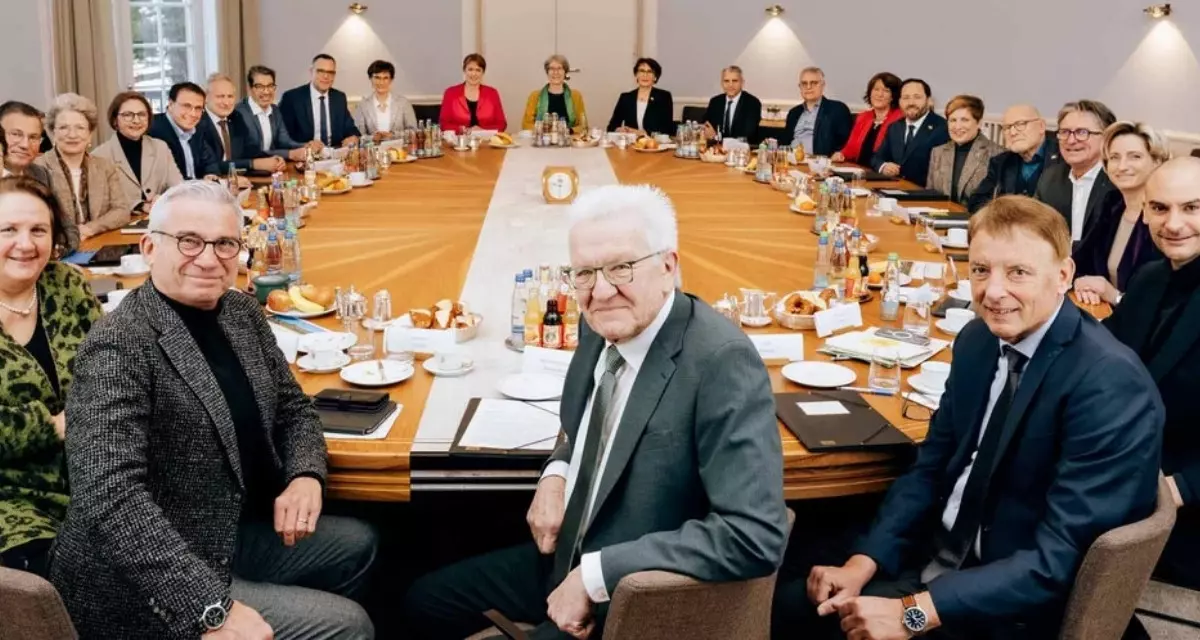 Alle Mitglieder des Kabinetts Baden-Württemberg sitzen an einem runden Tisch und schauen in die Kamera.