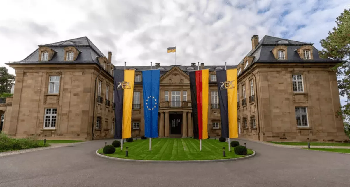 Das Staatsministerium Baden-Württemberg ist ein Sandsteingebäude in Front Ansicht mit vier Flaggen auf dem Hof gehisst: links und rechts außen die von Baden-Württemberg, in der Mitte die deutsche und europäische Flagge. 